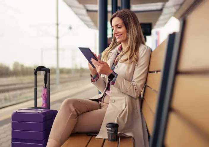 Une femme assise sur un banc attend son train une tablette à la main