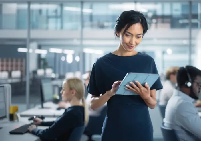 Une femme tient une tablette avec des techniciens en fond qui travaillent sur ordinateur